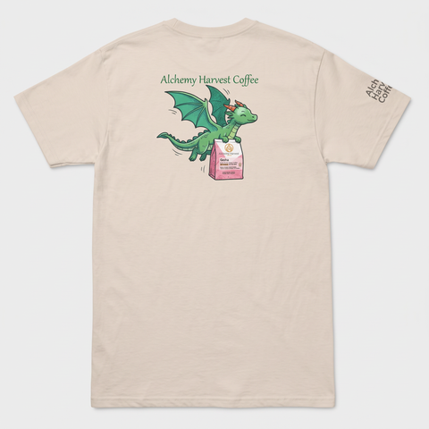 A light beige crewneck t-shirt shown from the back, featuring a cartoon green dragon holding a pink coffee bag with the text 'Alchemy Harvest Coffee' above it. The right sleeve has 'ALCHEMY.