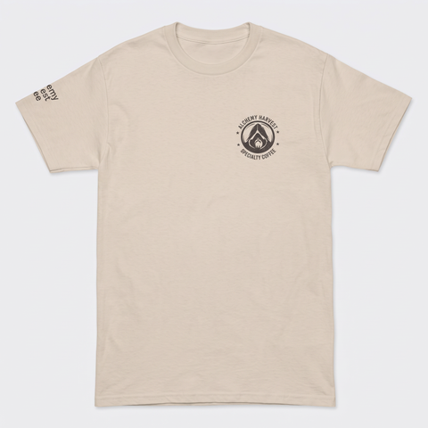 A light beige crewneck t-shirt with a black circular logo on the left chest reading 'ALCHEMY HARVEST COFFEE' and a small black text print on the right sleeve.