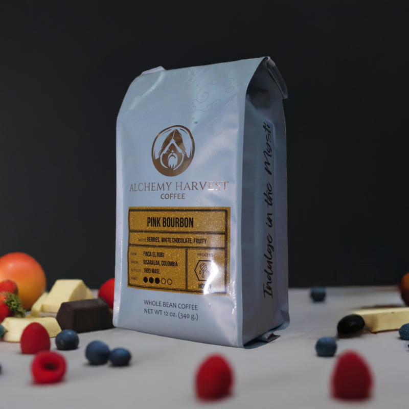A bag of Alchemy Harvest Coffee's Pink Bourbon honey process coffee beans, surrounded by an assortment of fruits and cheeses on a white surface.