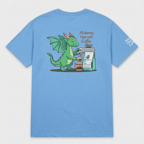 A light blue short-sleeve t-shirt with a cartoon graphic of a green dragon brewing coffee next to a coffee bag. The graphic includes the text 'Alchemy Harvest Coffee' and 'ALCHEMY HARVEST COFFEE'