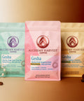 Three packages of Alchemy Harvest Coffee with flowers and coffee beans on a gradient background